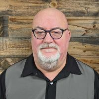 A bald man with glasses and a beard standing in front of a wooden wall.